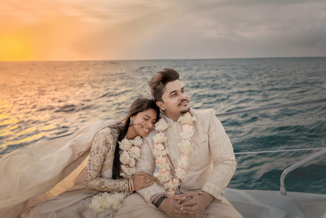 Destination yacht wedding in Dubai: couple in traditional cream sherwani and saree with floral garlands, seated together on yacht deck, golden sunset and Arabian Sea backdrop, ring ceremony moment