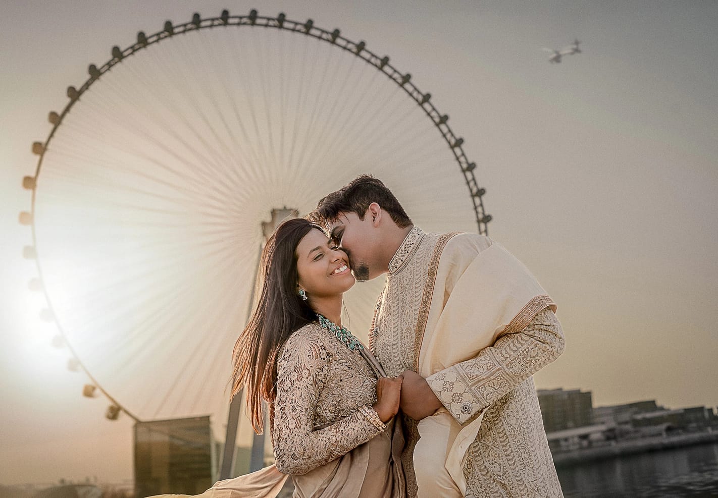 Wedding couple on yacht in Arabian Sea: bride in cream saree with floral garland, groom in cream sherwani, turquoise water and Ain Dubai hotel visible, destination wedding Dubai