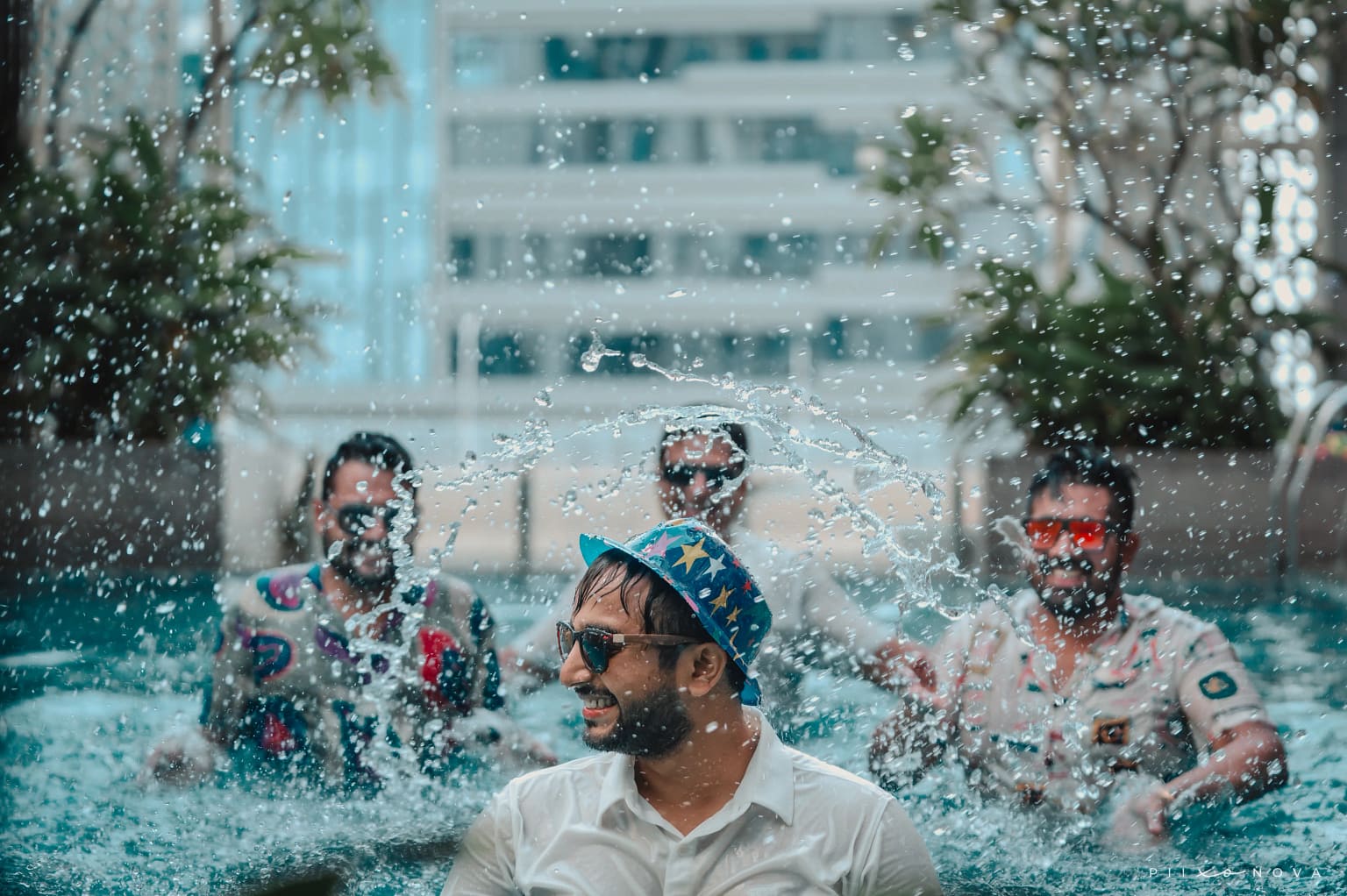 Group of men playing and splashing in swimming pool with water droplets flying, wearing sunglasses, bachelor party pool celebration, resort setting - wedding photography in Kolkata