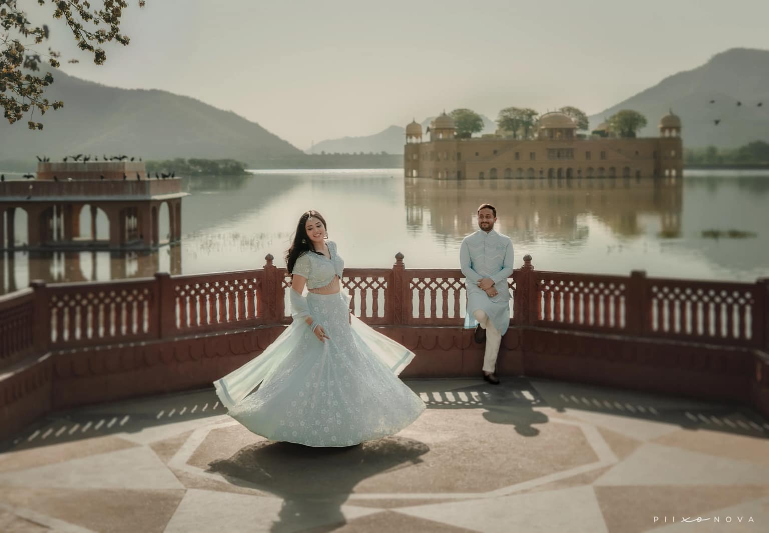 Bride twirling in flowing light-blue lehenga on red sandstone terrace, groom watching from railing, Jal Mahal water palace and lake backdrop, Jaipur destination pre-wedding shoot - pre-wedding shoot in Kolkata