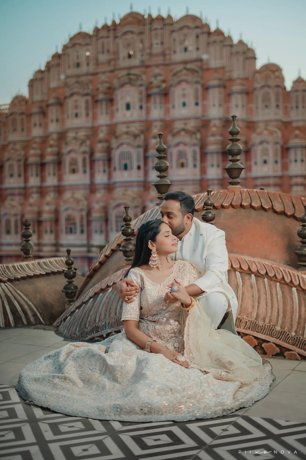 Groom tenderly kissing bride’s head as she sits in white embroidered lehenga, seated together on ornate rooftop, Hawa Mahal Palace of Winds in background, destination pre-wedding shoot, Jaipur - pre-wedding shoot in Kolkata