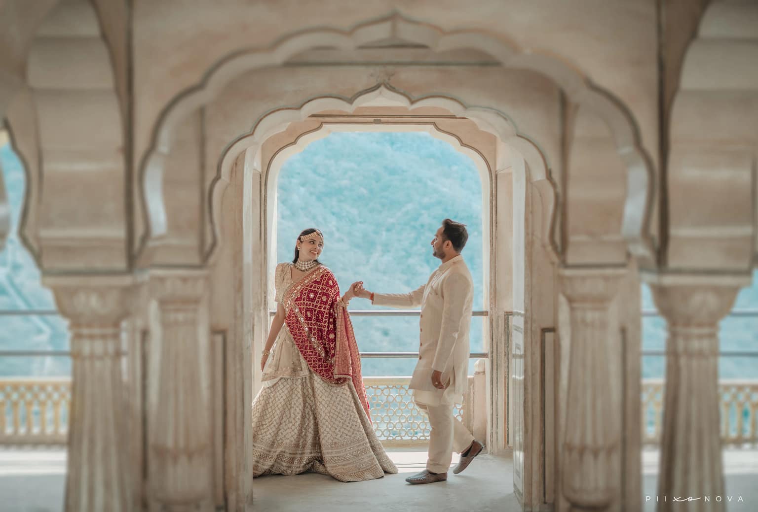 Couple holding hands framed by ornate scalloped palace archway with mountain vista, bride in cream lehenga with red dupatta, groom in cream kurta, destination pre-wedding shoot, Rajasthan - pre-wedding shoot in Kolkata
