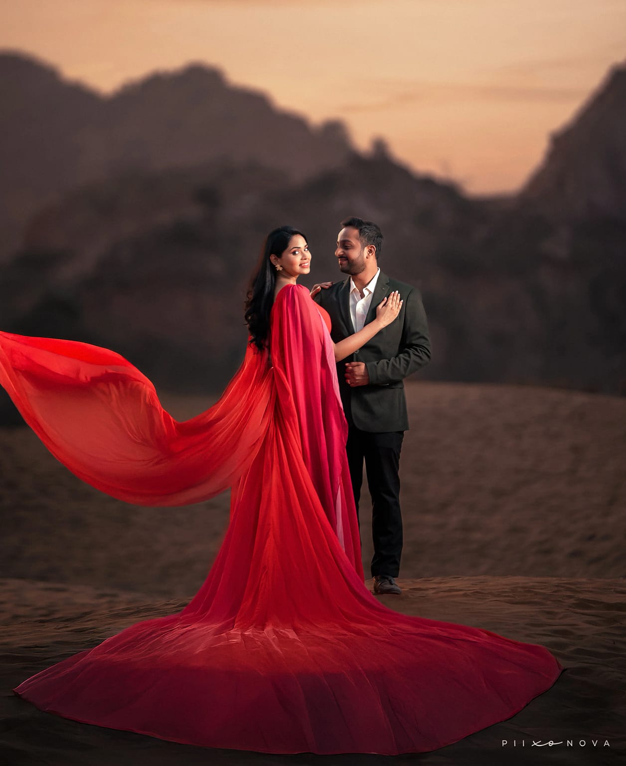 Couple in desert at dusk: woman in flowing red gown with dramatic wind-blown train, man in dark suit, mountain silhouettes and warm sunset sky, destination pre-wedding shoot - pre-wedding shoot in Kolkata
