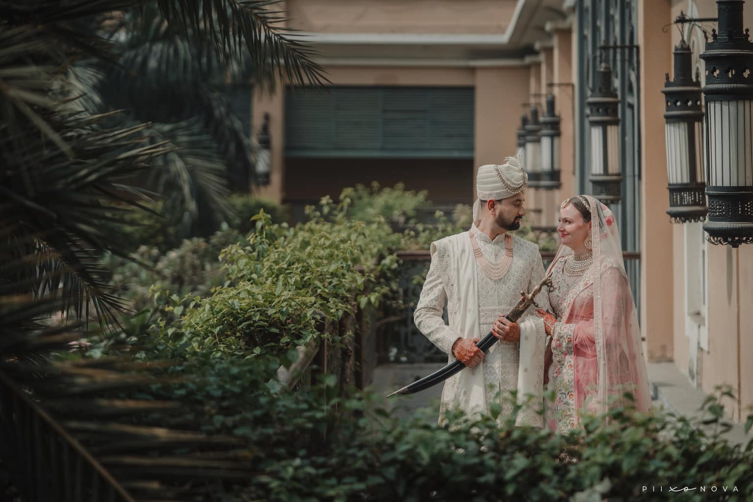 Groom holding ceremonial sword facing bride, traditional wedding ritual, groom in white turban and sherwani, bride in cream and pink veil, palm trees and heritage building backdrop, Kolkata