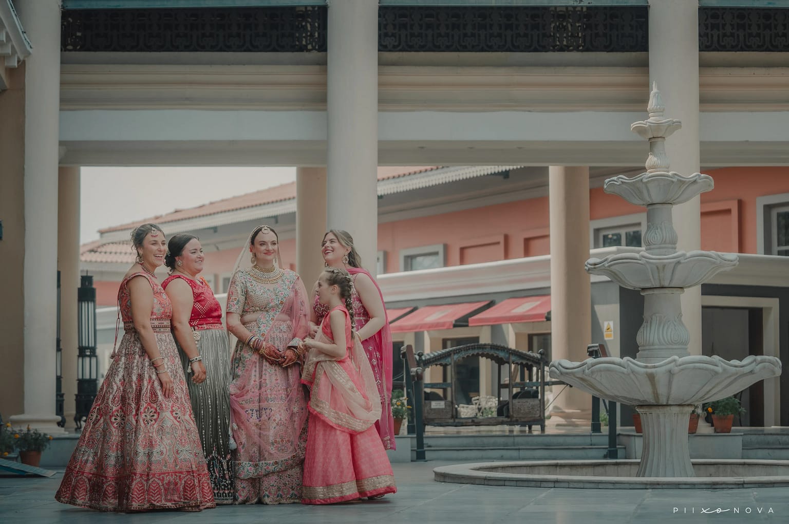 Bride surrounded by bridesmaids in coordinating pink and red outfits near ornate stone fountain, colonial-style columned pavilion, group wedding portrait, Kolkata