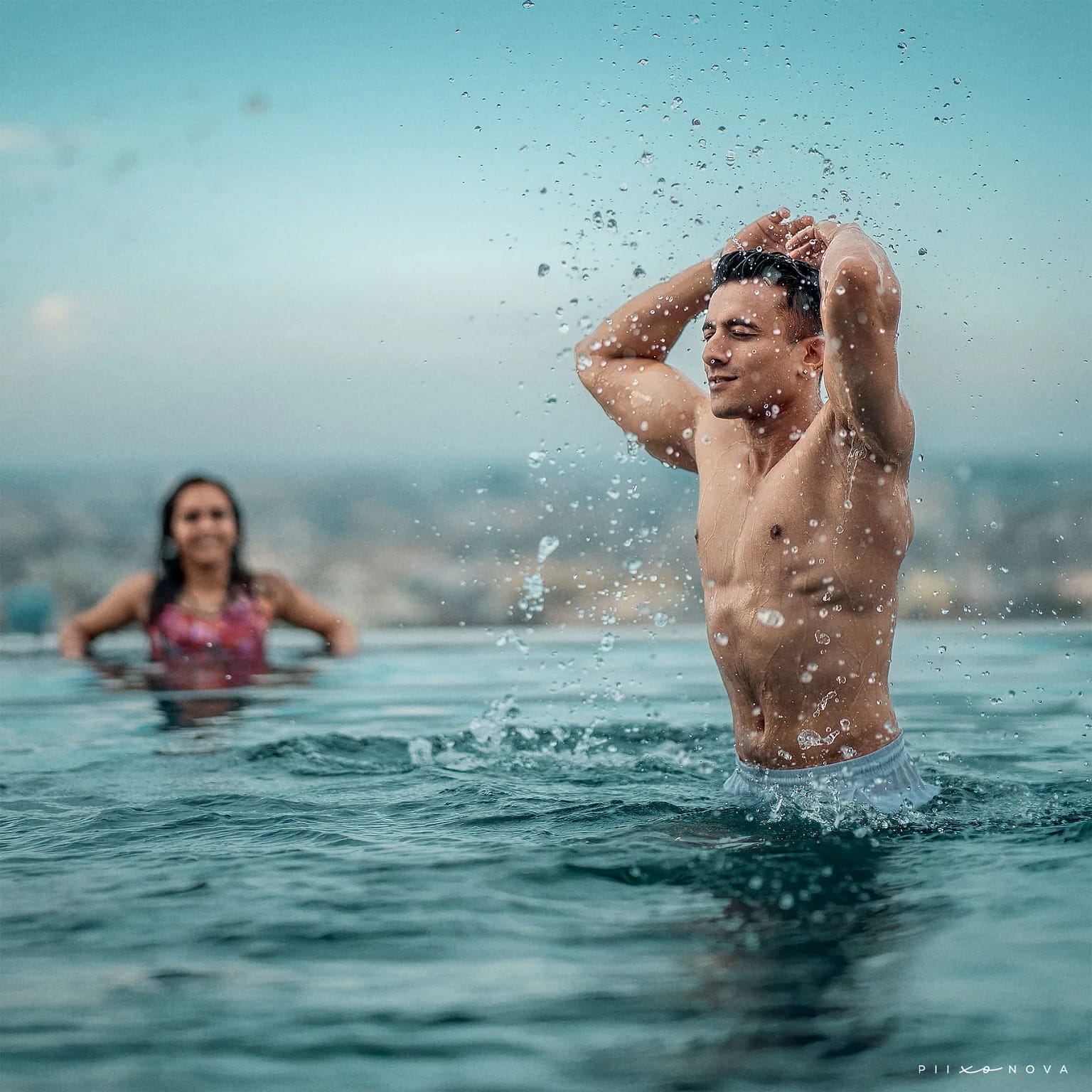 Man splashing water dramatically from his wet hair in infinity pool, woman smiling in blurred background, city skyline behind, playful resort pre-wedding shoot - pre-wedding shoot in Kolkata