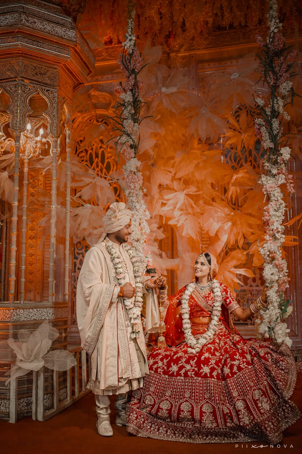 Bride seated on ornate floral-decorated swing wearing red embroidered lehenga with white garlands, groom standing beside her in cream sherwani and turban, richly decorated mandap stage, wedding ceremony, Kolkata - Bengali wedding photography in Kolkata
