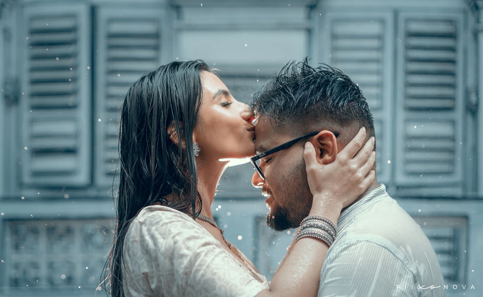 Couple embracing in the rain: woman kissing man's forehead tenderly, both soaking wet in casual white outfits, rustic wooden shutter background, romantic pre-wedding moment, Kolkata