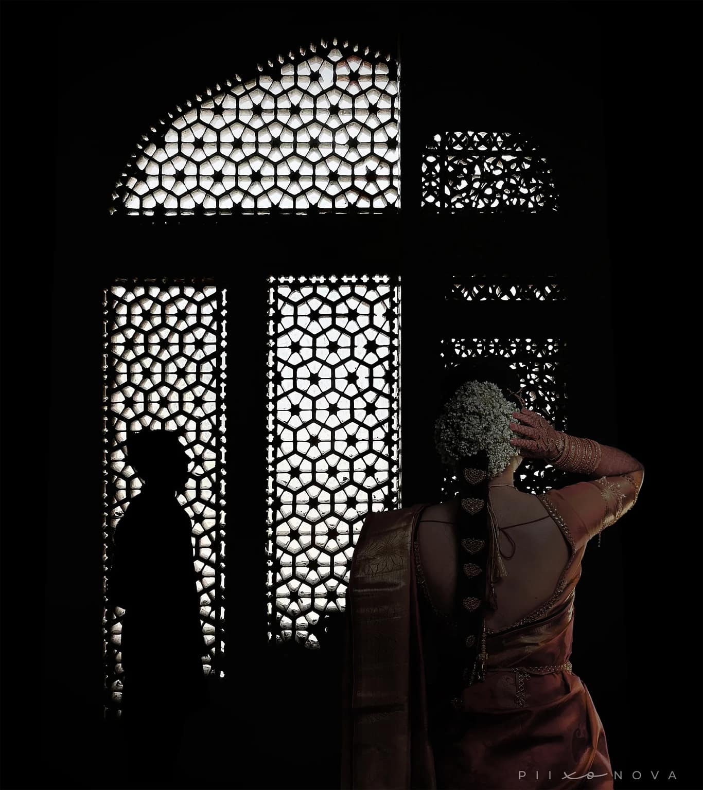 A woman stands gracefully in front of a beautifully designed jali window, showcasing intricate patterns, groom silhouette visible in the distance, artistic wedding portrait