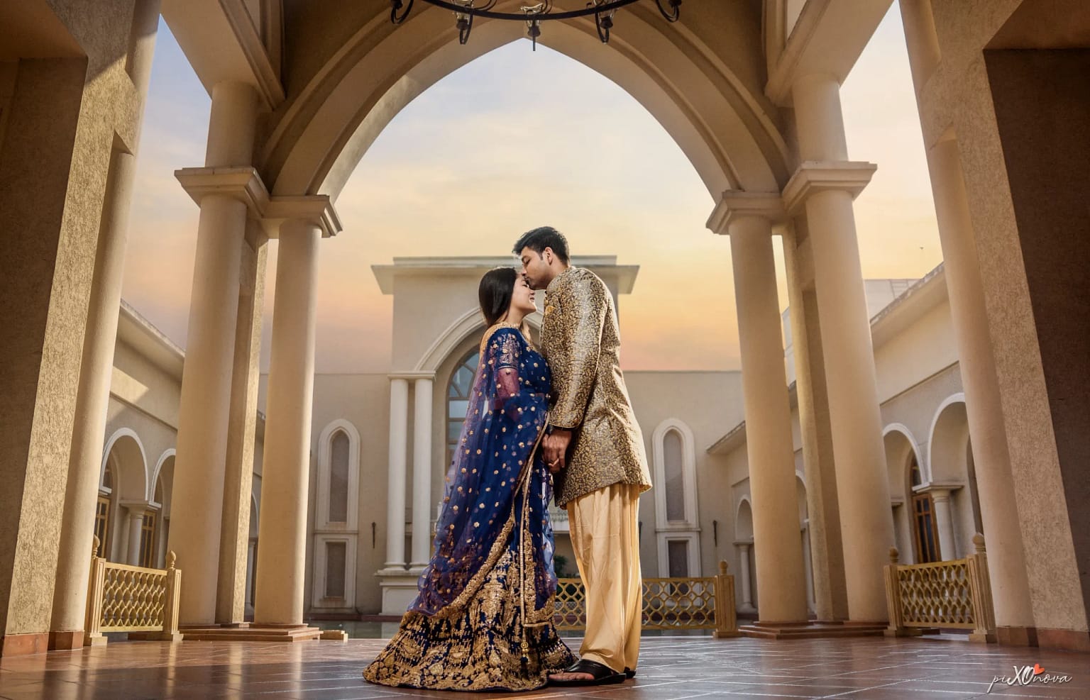 Couple in first dance moment in grand columned hall: bride in dark blue lehenga, groom in cream sherwani, golden hour wedding reception, Kolkata
