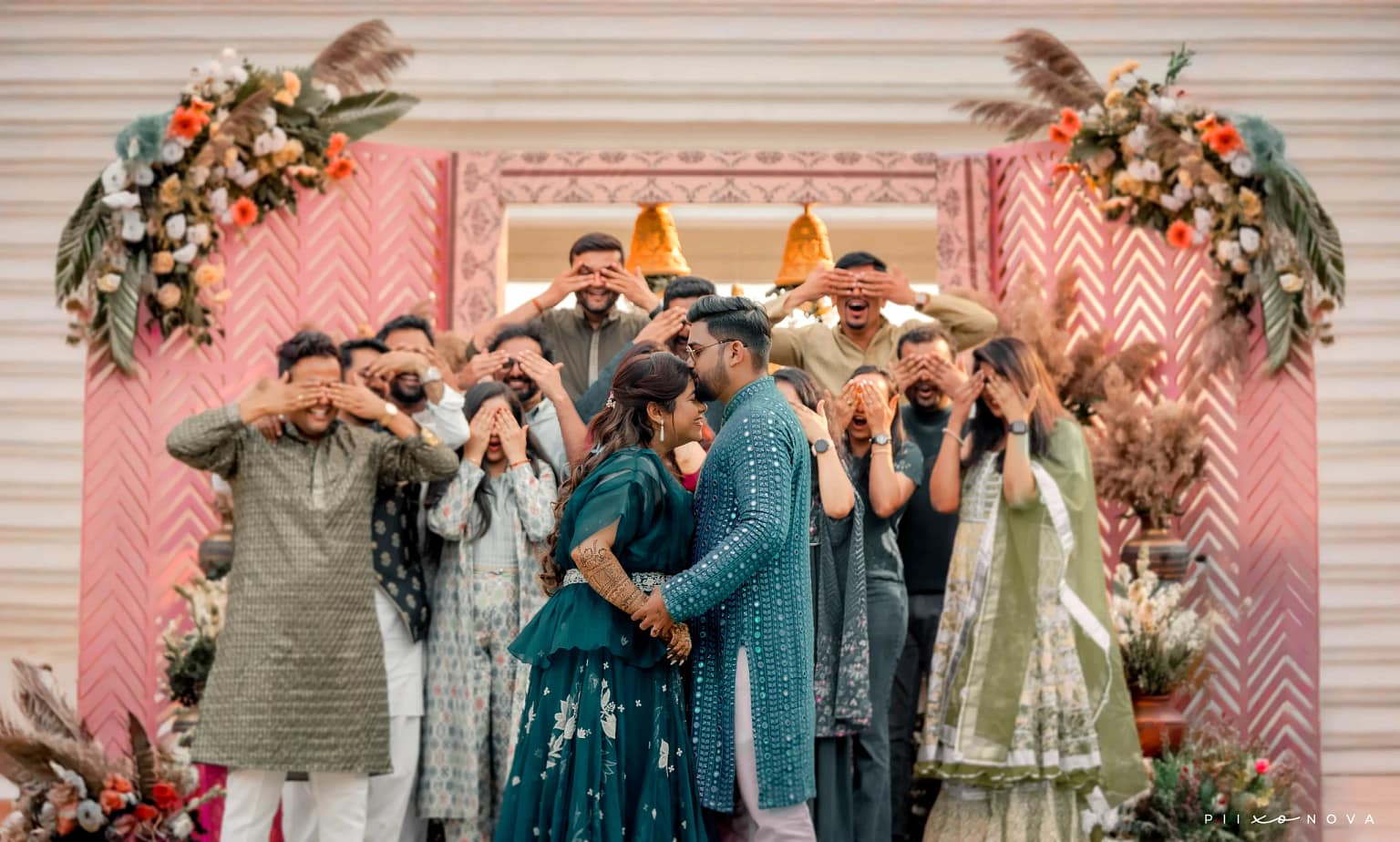 Ritika & Sumit in matching teal outfits about to kiss while guests playfully cover their eyes, pink floral chevron backdrop, fun candid wedding moment, Kolkata