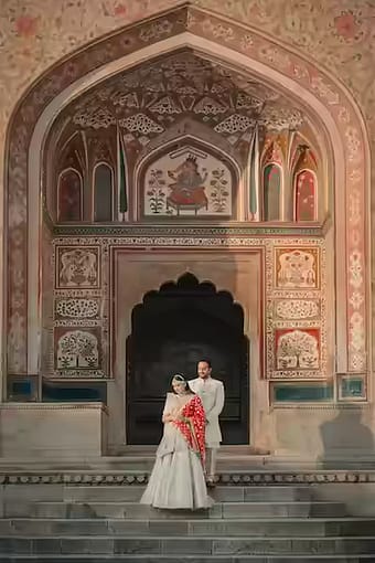 Couple standing beneath ornate painted archway with intricate frescoes, bride in white lehenga holding red decorative umbrella, groom in beige sherwani, Amer Fort-style palace architecture, destination pre-wedding shoot, Rajasthan - pre-wedding shoot in Kolkata