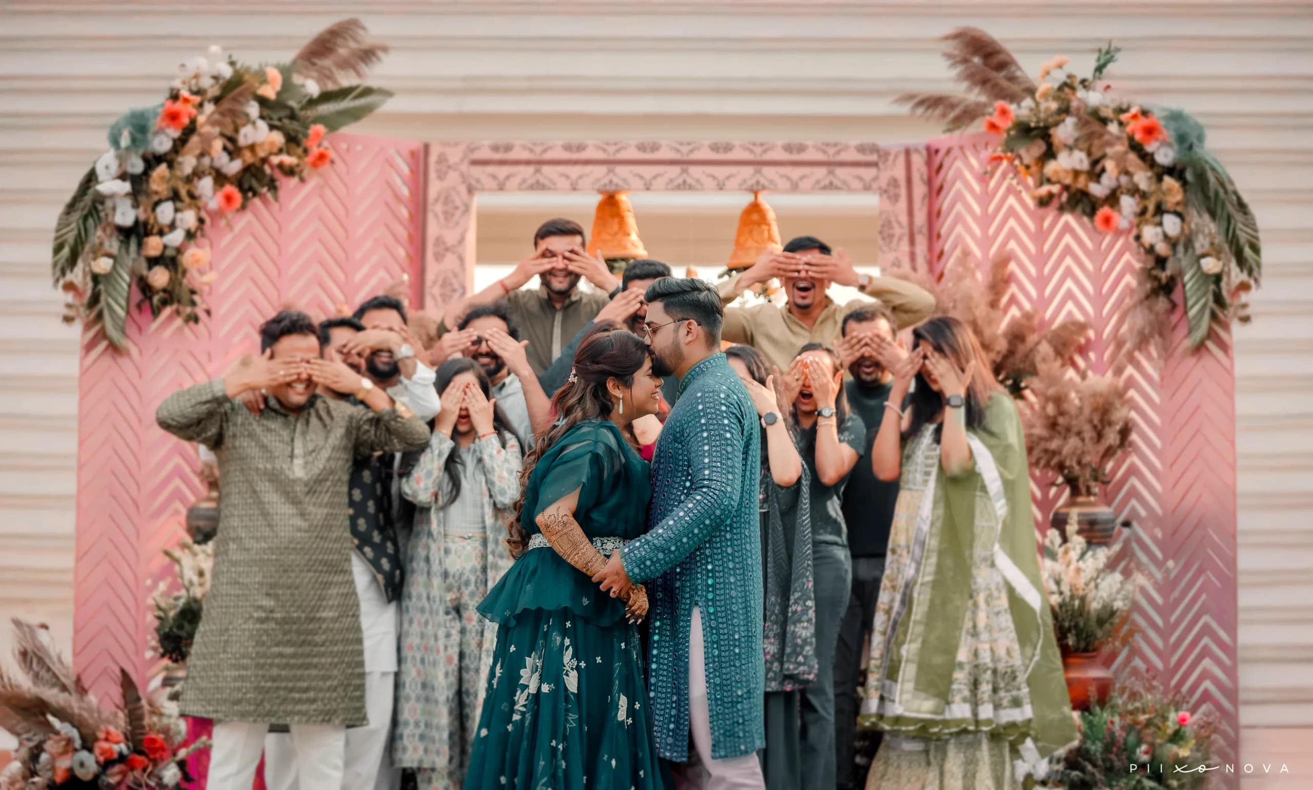 Ritika & Sumit in matching teal outfits about to kiss while guests playfully cover their eyes, pink floral chevron backdrop, fun candid wedding moment, Kolkata