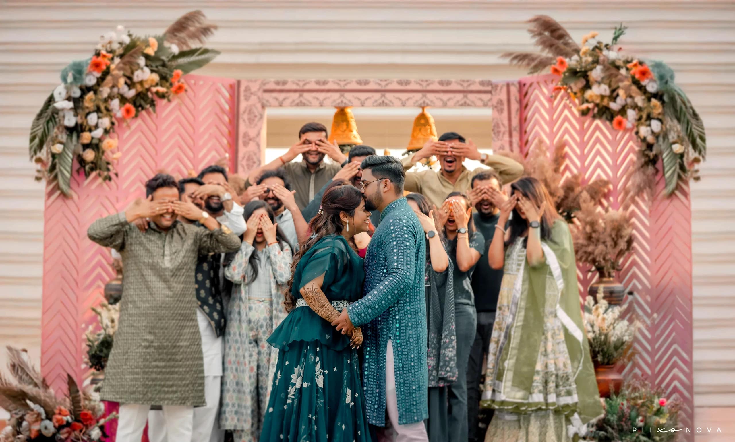 Ritika & Sumit in matching teal outfits about to kiss while guests playfully cover their eyes, pink floral chevron backdrop, fun candid wedding moment, Kolkata
