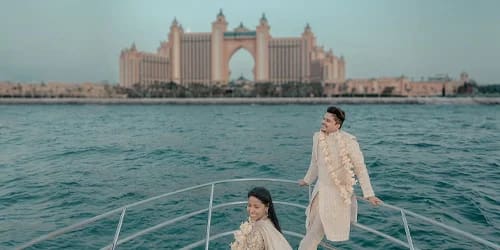 Destination yacht wedding: couple in cream traditional outfits with floral garlands standing on yacht bow, Atlantis The Palm hotel backdrop, Dubai