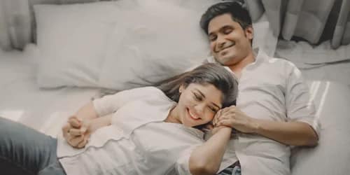Couple in intimate pre-wedding bedroom portrait: both in white t-shirts laughing together, cozy comfortable moment on bed with white pillows, Kolkata