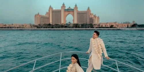 Destination yacht wedding: couple in cream traditional outfits with floral garlands standing on yacht bow, Atlantis The Palm hotel backdrop, Dubai