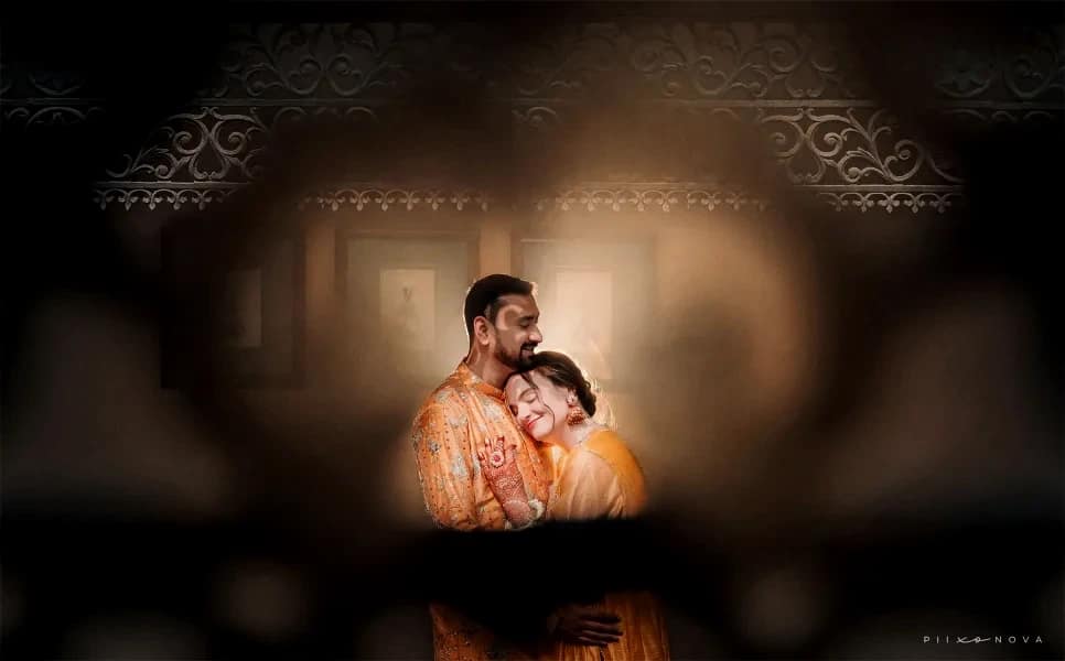 Couple embracing warmly in matching orange outfits, shot through blurred jali lattice foreground creating artistic peephole framing, warm intimate lighting, Kolkata - candid wedding photographer Kolkata